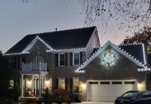Two-story house decorated with warm white LED Christmas lights along the roofline and gables.