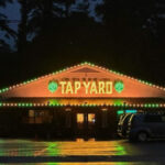 Exterior view of a bar with warm string lights running along the roofline and an illuminated "TAP YARD" sign at night.