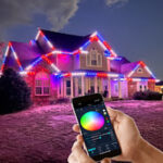 Hand using a smartphone app to change the color of permanent LED roofline lighting on a home at night