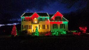Two-story house at night brightly illuminated with vibrant, multi-colored red, green, and yellow Christmas lights along the roofline and landscaping.