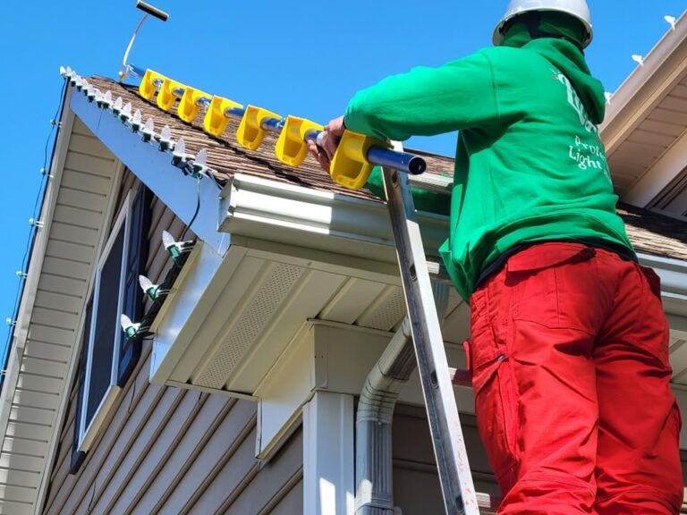 Professional installer on a ladder installing warm Christmas lights along a house roofline.