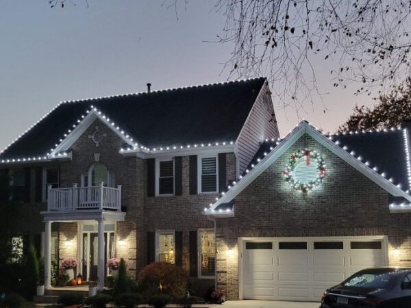 Two-story house decorated with warm white LED Christmas lights along the roofline and gables.