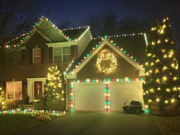 A holiday-decorated house featuring warm white roofline lights, wrapped porch pillars, and a lit Christmas tree in the front yard.